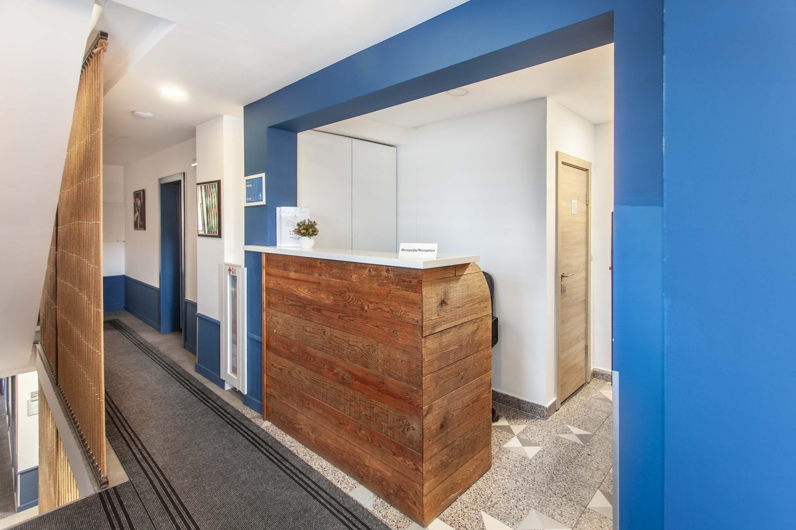 Dobrodošli u "Vilu Ars"! Fotografija prikazuje recepciju smeštaja, sa toplim drvenim pultom koji dominira prostorom. Plava boja na zidovima i iznad pulta stvara kontrast sa belim zidovima i drvenim elementima, dajući svež i moderan izgled.  Dugačak hodnik, sa tepihom i detaljima u plavoj boji, vodi do soba.  Pažljivo odabrani detalji, poput umetničkih slika i dekorativnih elemenata, doprinose prijatnoj atmosferi.  Sve je čisto, uredno i spremno da dočeka goste.